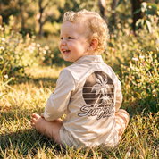 Child sitting in a grassy field wearing a 'Sunfolk Apparel' shirt with a palm tree design.
