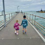 Two children holding hands on a pier with a cloudy sky and water in the background.