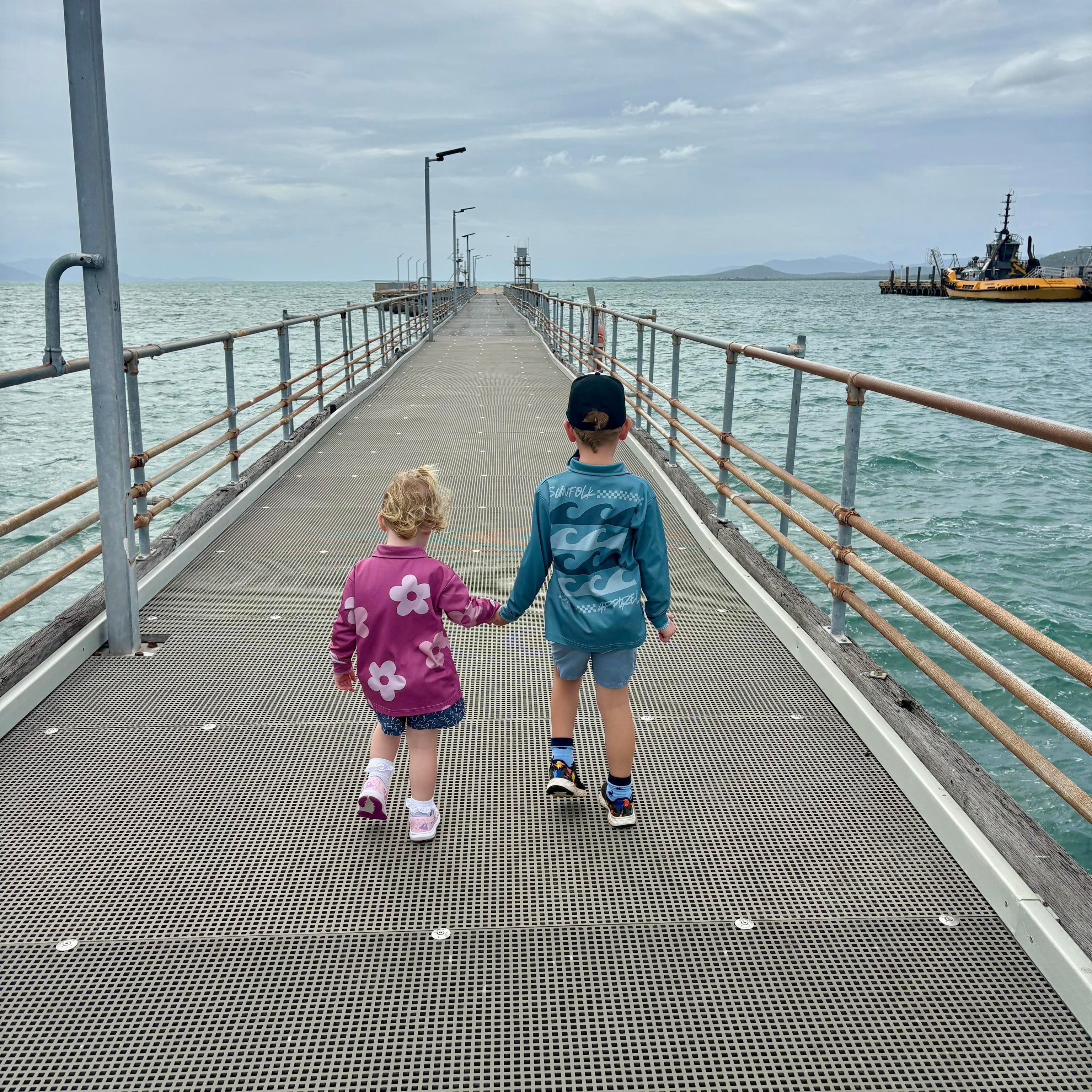 Two children holding hands on a pier with a cloudy sky and water in the background.