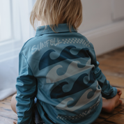 Child wearing a blue long sleeve sun safe shirt with wave designs sitting on a wooden floor.