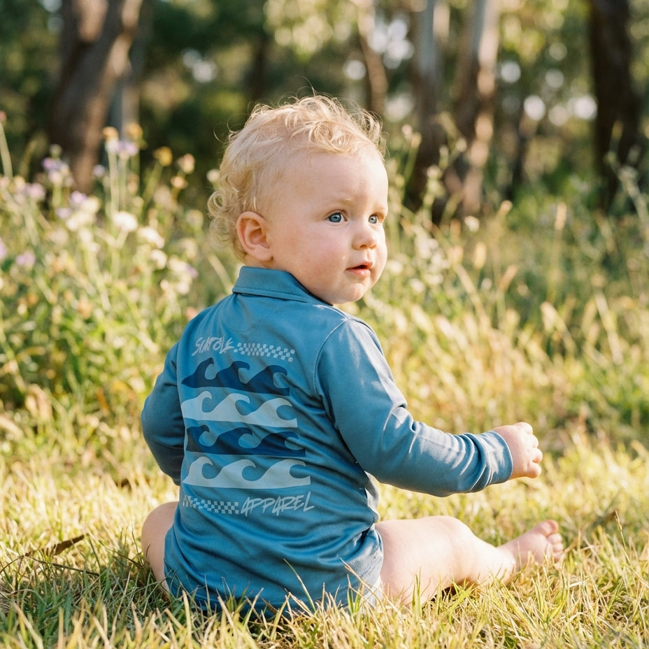 Baby sitting in a grassy field wearing a blue rash guard with wave patterns.